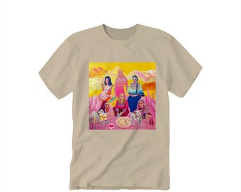 Op de afbeelding: Een beige t-shirt met een kleurrijke grafische print. Het kunstwerk toont een groep vrouwen in een landschap met roze en gele tinten. De vrouwen dragen verschillende outfits en sommigen houden dieren vast.