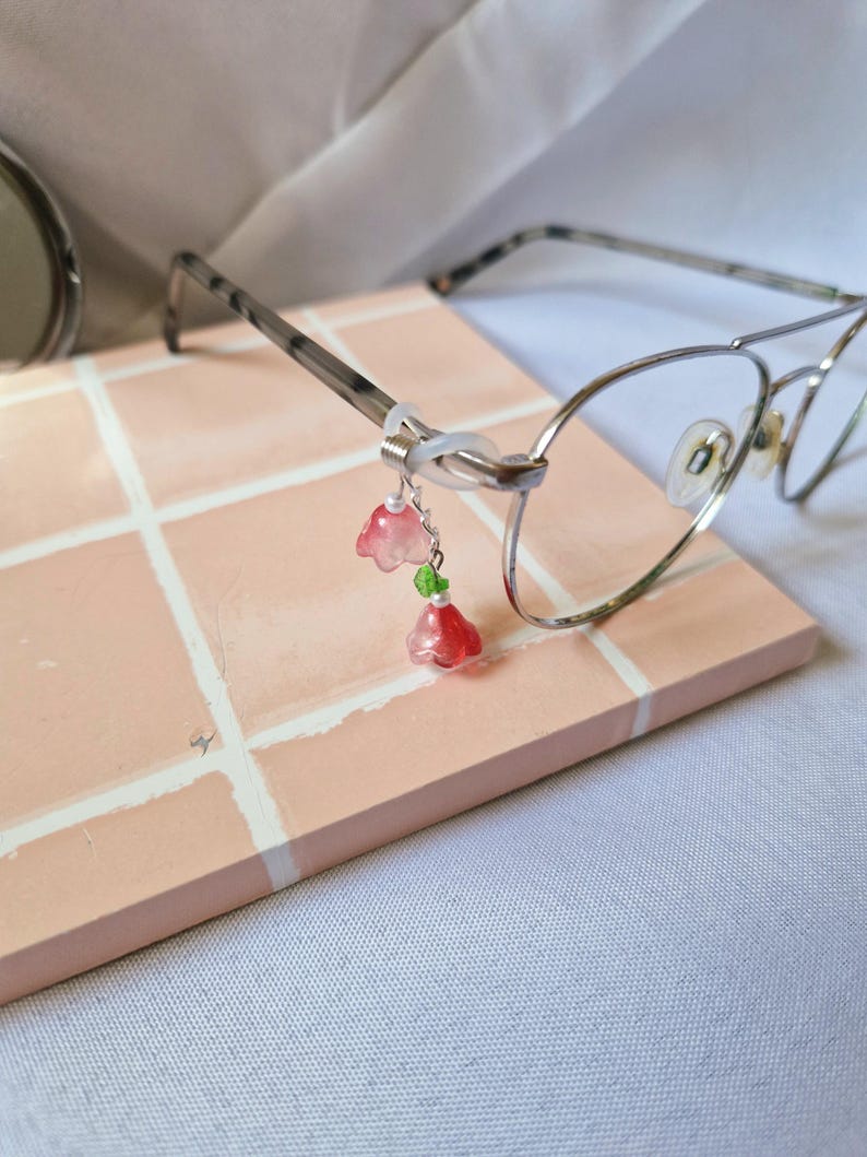 May include: A pair of silver-toned metal eyeglasses featuring a decorative charm attached to the temple. The charm consists of two translucent pink flower-shaped beads, a small green bead, and tiny white seed beads connected by silver wire.