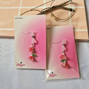 May include: Two pairs of dangle earrings featuring small red and pink flower charms with green leaves and white beads. The earrings are attached to pink gradient display cards with the text ukyo designs, and are encased in clear plastic sleeves.