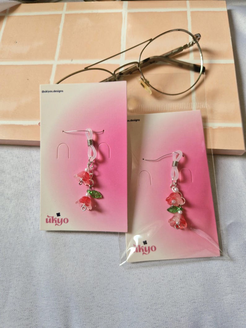 May include: Two pairs of dangle earrings featuring small red and pink flower charms with green leaves and white beads. The earrings are attached to pink gradient display cards with the text ukyo designs, and are encased in clear plastic sleeves.