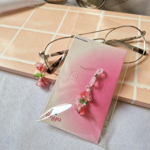 May include: A pair of eyeglass chains featuring small, translucent pink flower-shaped beads with green leaf accents and tiny white pearls. The chains are attached to clear silicone loops and are displayed on a pink gradient card labeled ukiyo.designs.