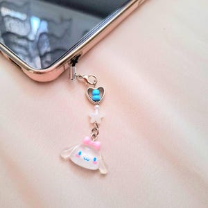 Puede incluir: Un charm de tel&eacute;fono con una cadena plateada, adjunto a un tel&eacute;fono de oro rosa. El charm presenta un coraz&oacute;n con cuentas azules, una estrella blanca y un personaje de dibujos animados transparente con un lazo rosa.