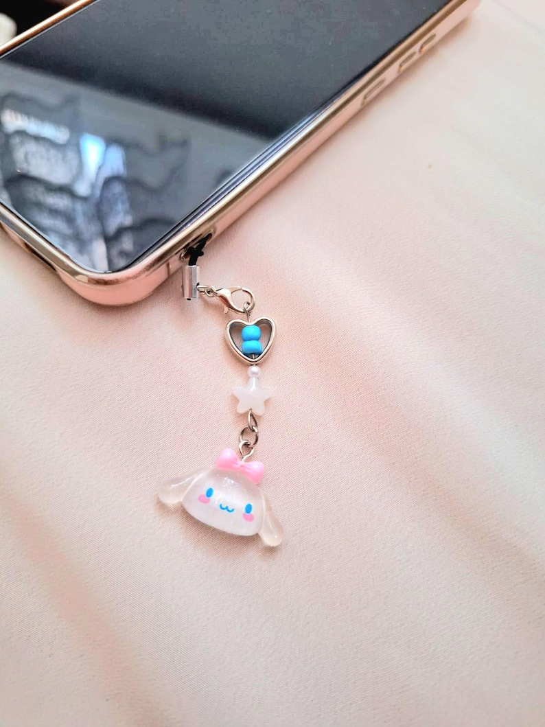 Puede incluir: Un charm de tel&eacute;fono con una cadena plateada, adjunto a un tel&eacute;fono de oro rosa. El charm presenta un coraz&oacute;n con cuentas azules, una estrella blanca y un personaje de dibujos animados transparente con un lazo rosa.