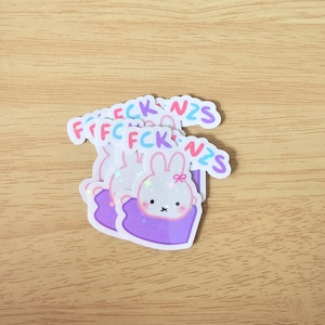 Puede incluir: Una pila de pegatinas troqueladas que muestran un conejo de dibujos animados blanco con mejillas rosadas y un pequeño lazo rosa, envuelto en una manta morada. Sobre el conejo, el texto FCK NZS está impreso en letras de colores.