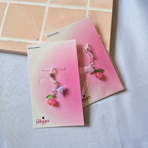 Puede incluir: Dos pares de dijes para gafas en tarjetas de presentación con degradado rosa etiquetadas como ukyo. Cada dije presenta una pequeña cuenta rosa, un acento de hoja verde y una pequeña flor unida a un lazo de silicona con un resorte de metal.