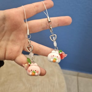 Könnte beinhalten: Zwei weiße und rosa Hasenanhänger mit Karottennasen und floralen Akzenten. Jeder Anhänger ist an einem silberfarbenen Metallverschluss mit einem herzförmigen Anhänger und einer weißen Kordel befestigt. Die Anhänger werden von einer Hand gehalten.