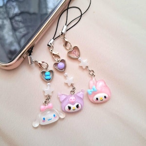 Puede incluir: Una colecci&oacute;n de bonitos colgantes para tel&eacute;fono con personajes de dibujos animados y cuentas en forma de coraz&oacute;n. Los colgantes son de colores pastel, incluyendo rosa, morado y blanco, y est&aacute;n sujetos a un tel&eacute;fono con un cord&oacute;n negro.
