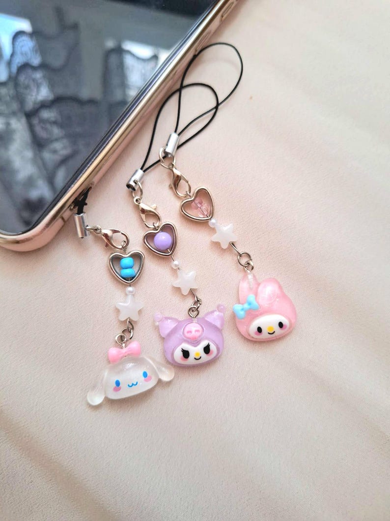 Puede incluir: Una colecci&oacute;n de bonitos colgantes para tel&eacute;fono con personajes de dibujos animados y cuentas en forma de coraz&oacute;n. Los colgantes son de colores pastel, incluyendo rosa, morado y blanco, y est&aacute;n sujetos a un tel&eacute;fono con un cord&oacute;n negro.