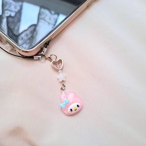 Puede incluir: Un charm de tel&eacute;fono rosa con un conejito de dibujos animados con un lazo azul, un charm en forma de coraz&oacute;n y cuentas en forma de estrella. El charm est&aacute; sujeto a un smartphone.