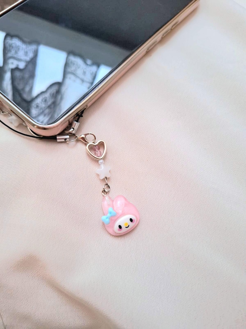 Puede incluir: Un charm de tel&eacute;fono rosa con un conejito de dibujos animados con un lazo azul, un charm en forma de coraz&oacute;n y cuentas en forma de estrella. El charm est&aacute; sujeto a un smartphone.