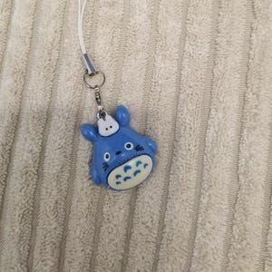 May include: A blue Totoro-shaped charm with a white hat and belly, attached to a silver-colored metal clasp and a white lanyard. The charm is lying on a textured, light-colored surface.