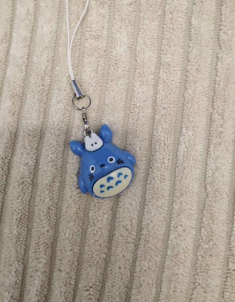 May include: A blue Totoro-shaped charm with a white hat and belly, attached to a silver-colored metal clasp and a white lanyard. The charm is lying on a textured, light-colored surface.