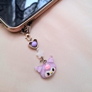 Puede incluir: Un charm de tel&eacute;fono con un coraz&oacute;n morado, una estrella blanca y un personaje de dibujos animados lavanda con una nariz rosa y ojos negros. El charm est&aacute; sujeto a un tel&eacute;fono de color dorado.