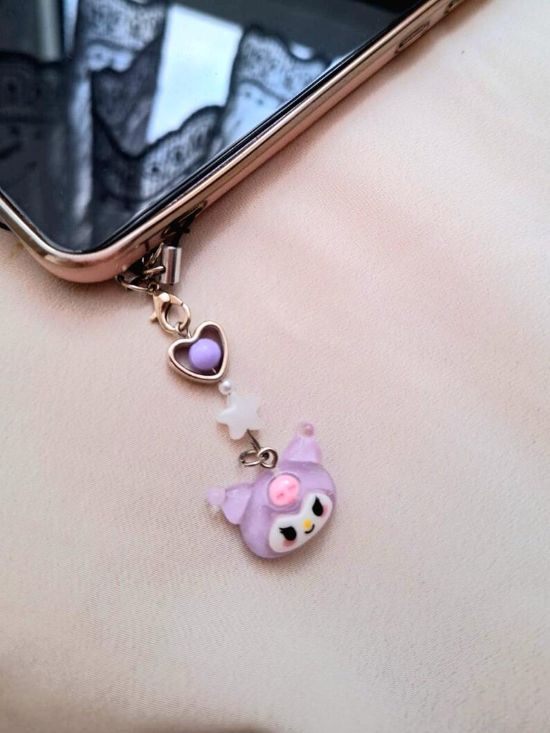 Puede incluir: Un charm de tel&eacute;fono con un coraz&oacute;n morado, una estrella blanca y un personaje de dibujos animados lavanda con una nariz rosa y ojos negros. El charm est&aacute; sujeto a un tel&eacute;fono de color dorado.