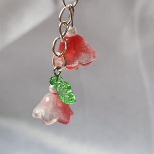 May include: A dangling charm featuring two bell-shaped flower beads in shades of red and pink, accented with small white spherical beads and a single textured green leaf bead, all suspended from a silver-toned metal chain and a translucent ring.