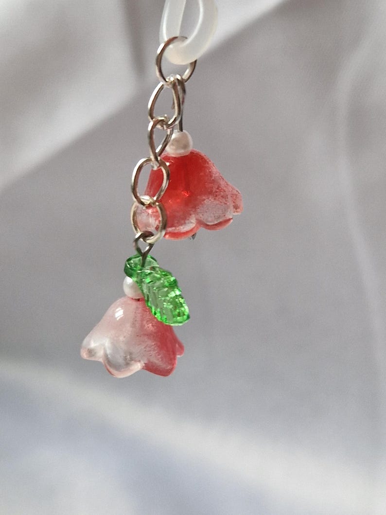 May include: A dangling charm featuring two bell-shaped flower beads in shades of red and pink, accented with small white spherical beads and a single textured green leaf bead, all suspended from a silver-toned metal chain and a translucent ring.