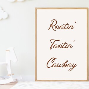May include: A framed print with the words "Rootin' Tootin' Cowboy" in brown script on an off-white background. The frame is light brown. The print is in a child's room with a white rocking horse and cloud decorations.