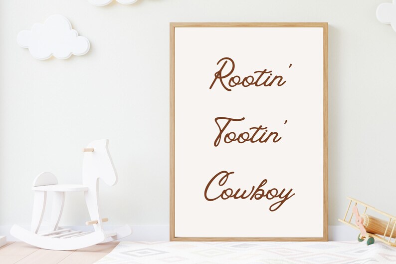 May include: A framed print with the words "Rootin' Tootin' Cowboy" in brown script on an off-white background. The frame is light brown. The print is in a child's room with a white rocking horse and cloud decorations.