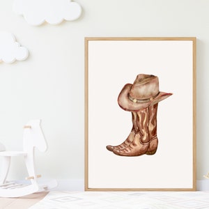 May include: Watercolor illustration of a cowboy boot and hat. The brown boot and hat are set against a white background, framed in a light wood. The artwork evokes a rustic, Western theme, suitable for nursery decor.