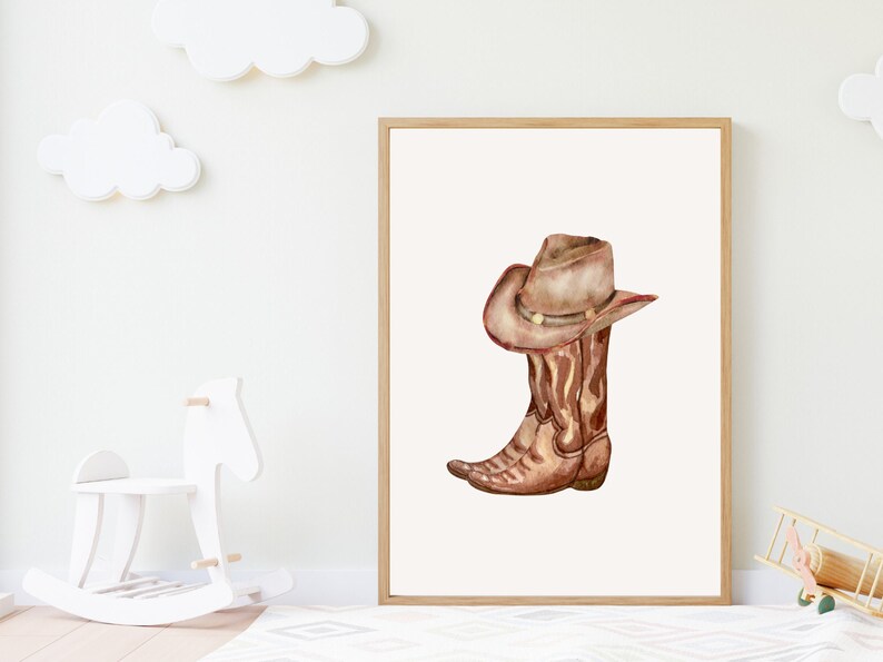 May include: Watercolor illustration of a cowboy boot and hat. The brown boot and hat are set against a white background, framed in a light wood. The artwork evokes a rustic, Western theme, suitable for nursery decor.