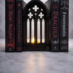 Könnte beinhalten: Ein schwarzer Kerzenhalter im gotischen Stil in Fensterform, flankiert von vier Büchern. Die schwarzen Bücher tragen Titel wie "Dracula" und "Frankenstein" in roter und weißer Schrift. Eine brennende Kerze ist im Halter sichtbar.
