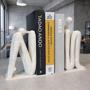 Può includere: Reggilibri astratti bianchi a forma umana seduta, che sorreggono tre libri. I libri sono neri, gialli e grigi, con titoli come "Tadao Ando" e "Mies". I reggilibri sono su una mensola bianca.