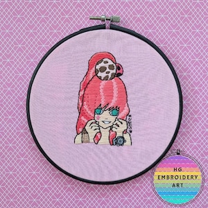 Puede incluir: Arte de bordado cosido a mano que presenta a una chica de dibujos animados con cabello rosado, ojos verdes y un accesorio manchado marrón y blanco. El bordado está en un aro negro sobre un fondo de tela rosa. El texto "*smile*" está incluido.