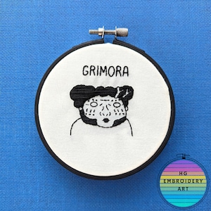 以下が含まれることがあります： 黒い刺繍で「Grimora」という文字が書かれた白い刺繍枠に、漫画のキャラクターが刺繍されています。