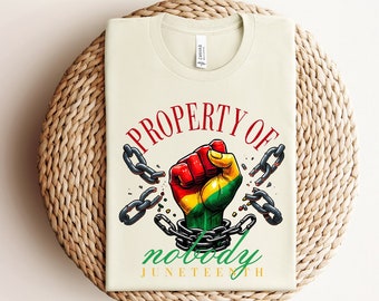 May include: A beige t-shirt with the text "PROPERTY OF nobody JUNETEENTH" in red and green. A black fist breaking free from chains is in the center of the shirt.