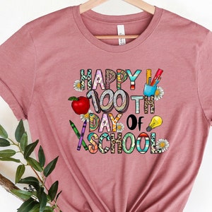 Puede incluir: Una camiseta color malva con el texto "Happy 100th Day of School" en letras de colores decoradas con &uacute;tiles escolares y flores.