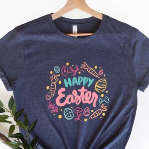 May include: A navy blue t-shirt with a colorful graphic design that says "Happy Easter" surrounded by images of carrots, eggs, and flowers.