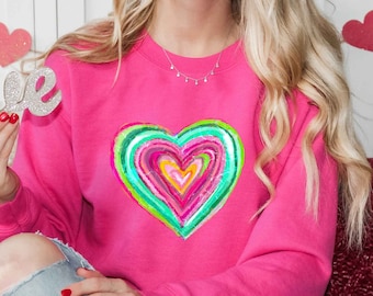 May include: Pink sweatshirt with a colorful, multi-layered heart design. The word "love" is written in silver glitter.