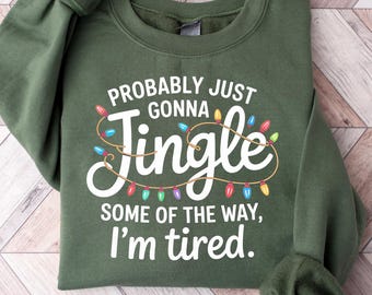 May include: An olive green sweatshirt with the text "Probably Just Gonna Jingle Some of the Way, I'm tired." The text is in white with a string of colourful Christmas lights. The sweatshirt is folded on a light wood surface.