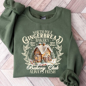 May include: Green sweatshirt with a Christmas-themed design featuring a gingerbread house and the text "North Pole Gingerbread Bakery. Merry Xmas. Warm Wishes. Bakery Club. Always Fresh."