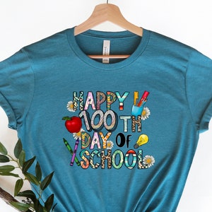 Puede incluir: Camiseta azul turquesa con un dise&ntilde;o gr&aacute;fico que dice "Happy 100th Day of School". El gr&aacute;fico est&aacute; formado por &uacute;tiles escolares coloridos como l&aacute;pices de colores, l&aacute;pices, una manzana y una bombilla.