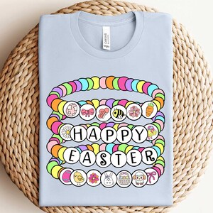 May include: Light blue t-shirt with a colorful beaded design that spells out "Happy Easter". The beads are decorated with small Easter-themed images.