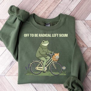 May include: Olive green sweatshirt with the text "OFF TO BE RADICAL LEFT SCUM." The graphic depicts a frog riding a bicycle with a cat in the basket. The bicycle is cream-colored with black tires.