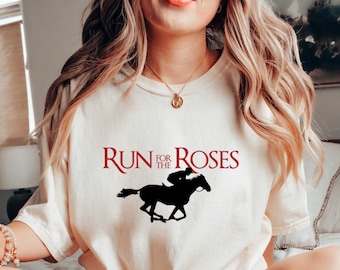 May include: A white t-shirt with the text "RUN FOR THE ROSES" in red lettering and a black silhouette of a jockey riding a horse. The shirt is being worn by a person sitting on a bed with a book and a mug.