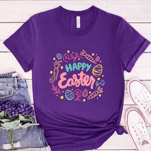 May include: Purple t-shirt with a white crew neck and a colorful graphic design that says "Happy Easter" surrounded by carrots, eggs, and flowers.