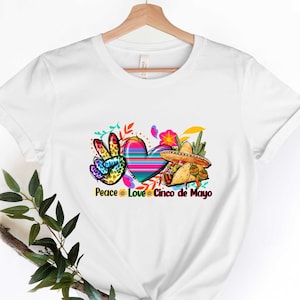 Puede incluir: Camiseta blanca con un dise&ntilde;o colorido de signo de paz, coraz&oacute;n y sombrero. El texto "Peace Love Cinco de Mayo" est&aacute; impreso debajo del dise&ntilde;o.
