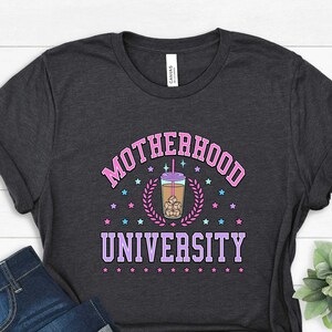 May include: A dark gray t-shirt with the text "MOTHERHOOD UNIVERSITY" in pink and purple lettering. A cartoon illustration of a cup of iced coffee with a straw and ice cubes is in the center of the text. The text is surrounded by small pink and blue stars.