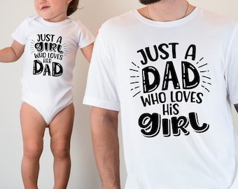Puede incluir: Una camiseta blanca con texto negro que dice "Just a Dad who loves his Girl". Un body blanco con texto negro que dice "Just a Girl who loves her Dad" es usado por un niño de pie sobre una mesa.