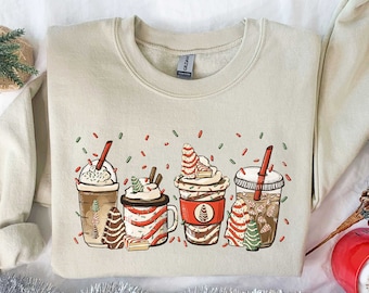 May include: A beige sweatshirt with a festive design of four different holiday-themed coffee drinks. The drinks are decorated with red and green sprinkles, candy canes, and Christmas trees.
