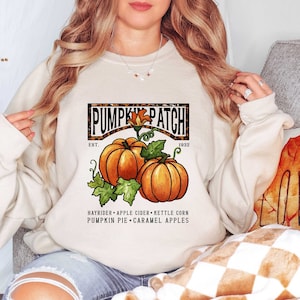 May include: A beige sweatshirt with a graphic print of a pumpkin patch. The graphic features a brown and black leopard print border with the text "Pumpkin Patch" in white. The text "Est. 1932" is printed below the graphic. The graphic also includes two pumpkins with green leaves and the text "Hayrider - Apple Cider - Kettle Corn Pumpkin Pie - Caramel Apples".