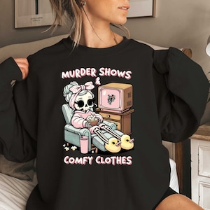 May include: Black sweatshirt with a graphic of a skeleton wearing a pink robe and watching TV. The text on the sweatshirt reads "Murder Shows & Comfy Clothes".