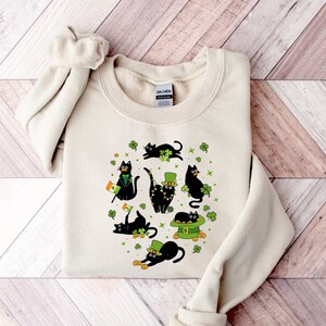 May include: A cream-colored sweatshirt with a black cat design. The cats are wearing shamrocks and leprechaun hats. The design is surrounded by green shamrocks.