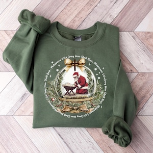 May include: Green sweatshirt featuring a Christmas-themed graphic. The graphic shows Santa Claus kneeling before a manger inside a snow globe, with text around the globe. The sweatshirt has long sleeves and a crew neck.