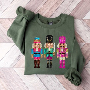 May include: A green sweatshirt with a festive design of three nutcracker soldiers. The nutcrackers are decorated with glitter and have colorful accents.