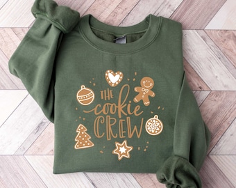 May include: A green sweatshirt with the text "The cookie crew" in brown lettering. The sweatshirt has a gingerbread man, a heart, a Christmas tree, a star, and an ornament graphic.