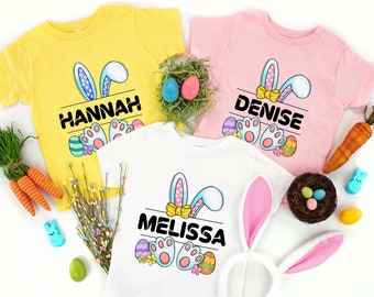 May include: Three white t-shirts featuring a bunny rabbit design with the names "Hannah", "Denise", and "Melissa" printed on them. The shirts are surrounded by Easter decorations, including carrots, eggs, and a nest.